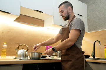 A man in a brown apron cooks at the stove in a sleek kitchen. He stirs a saucepan and organizes...