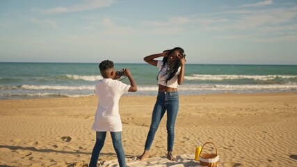 Happy african american siblings capturing memories with classic camera as they have fun on beach, with sister dancing and striking poses during sunny seaside picnic
