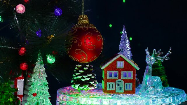 On a dark blue background with laser animation, near a red ball with a pattern hanging on a large decorated Christmas tree branch, an orange house surrounded by a pine cone and a deer with Christmas t