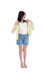 Young, barefoot Asian girl putting on light yellow cardigan over white tank top and denim shorts, standing against white background.