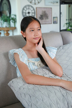 Portrait young girl sitting comfortably on sofa, resting her hand on her cheek and looking contemplatively to the side in a cozy home interior.