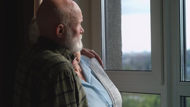 Caring senior man hugging and consoling sad wife near window, emotional support