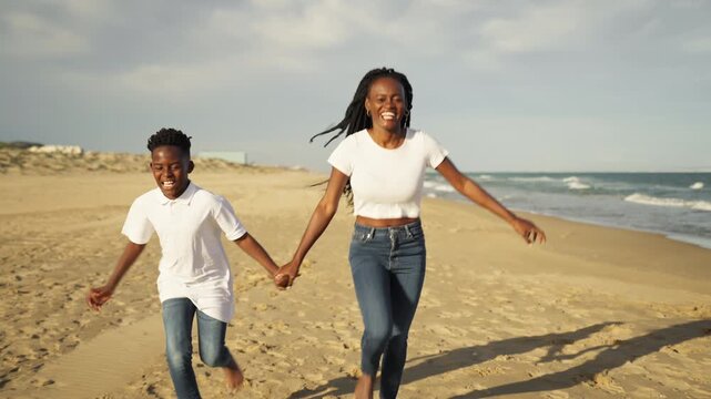 Happy african american mother and her cheerful son holding hands and running together barefoot on sandy beach, enjoy beautiful sunny day and their family summer vacation by sea