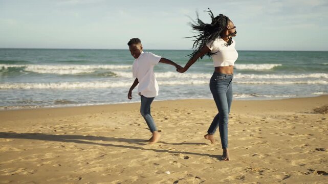 Happy african american mother and her son holding hands and joyfully running and dancing barefoot on sandy beach during beautiful sunny day, enjoying their family vacation together