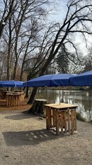 Calm lakeside park at the Christmas market in Bad Aibling, featuring blue umbrellas, wooden tables, festive decorations and a peaceful atmosphere surrounded by trees and seasonal charm.