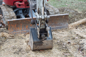 Close-up of a red mini excavator bucket and dozer blade resting on straw-covered ground at a job site