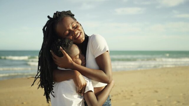 Loving african american mother with dreadlocks tenderly embracing young son on sandy beach, sharing warm and happy moment together with serene ocean waves in background