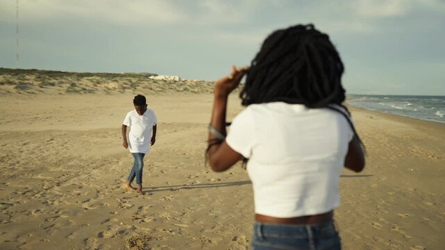 Back view of young mother with braids recording ittle boy happily dancing barefoot on sandy beach during beautiful sunny day, creating cherished family memories together
