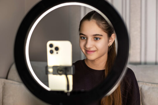 Teen girl smiling while filming with phone. A happy teenage girl sits behind ring light, smiling at the smartphone camera while recording video content at home for social media.