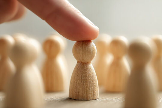 Finger selecting one wooden figurine among many as a symbol of leadership decision making and individual choice in a group setting with blurred background figures