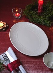Christmas table setting on a burgundy tablecloth with a white oval platter, candles, and a napkin.