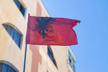 Albanian red and black national flag waving in city street, building and sky background. Symbol of patriotism, pride, tourism, and travel in Europe