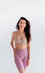 Fototapeta premium Sports woman standing in activewear with abs muscles, looking at camera isolated on white background. Caucasian 30s fitness girl wears beige top, pink leggings. Sexy fit smiling brunette, blue eyes.