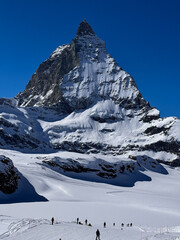 Winter view of the Matterhorn rising above a snowy glacier with small skiers below. Clear blue sky, alpine landscape, and dramatic mountain scenery in Zermatt, Switzerland.