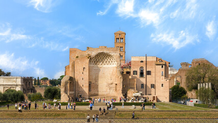 View of Santa Francesca Romana Basilica in the Roman Forum, Rome, Italy, with ancient ruins, a Romanesque bell tower, and crowds of visitors under a bright blue sky.