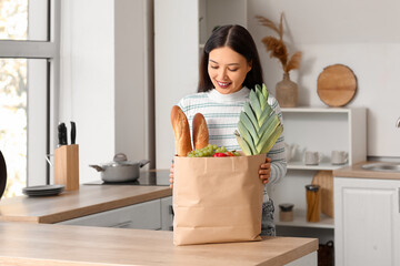 Young Asian woman with shopping bag full of fresh food at table in kitchen