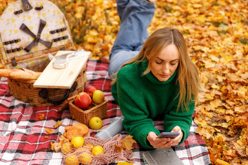 Mid-adult woman lying prone on a red plaid blanket covered in fall leaves, looking at her smartphone during a picnic. Combining outdoor relaxation with connectivity.