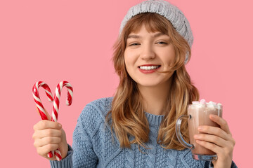 Pretty young woman with cup of hot chocolate and candy canes on pink background