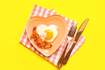 Plate with tasty bacon, heart made of fried egg and cutlery on yellow background