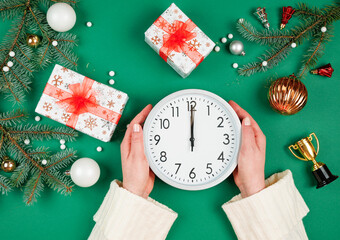 Female hands holding a round clock marking Christmas Eve midnight. Set to 12 AM. Festive time New Year concept with ornaments and gifts on green. First person top view photo. 