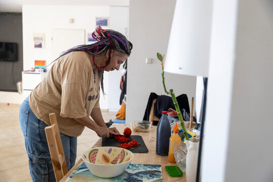Plus size woman with dreadlocks standing at a table, concentrated on slicing a tomato with a knife on a cutting board while preparing food. - Powered by Adobe