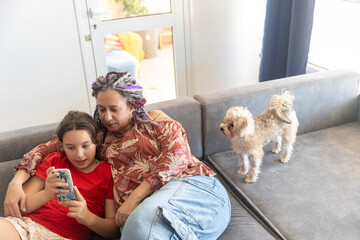Plus size woman with dreadlocks and a young girl sitting together on a sofa, looking at a smartphone screen.