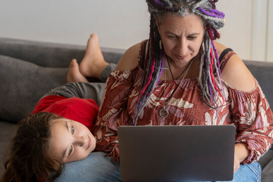 Plus size woman with dreadlocks working on a laptop while a young girl is resting and leaning on her lap on the sofa. - Powered by Adobe