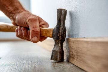 Handheld hammer with wooden handle being used to install baseboard trim along a wall during home interior renovation or flooring project