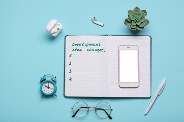 Open notebook with text DEVELOPMENT IDEA CONCEPT, mobile phone and alarm clock on blue background. Top view