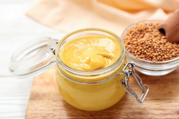 Board with jar of mustard and bowl of seeds on white wooden background