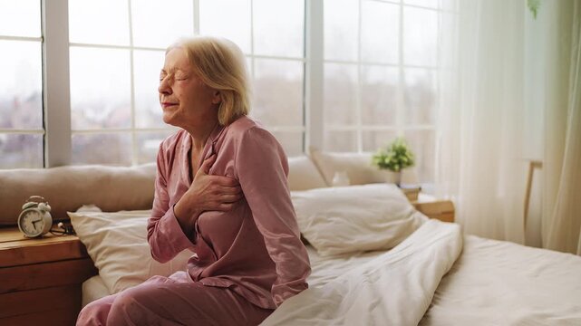 Stressed senior woman holding chest in pain while sitting on bed, heart risk
