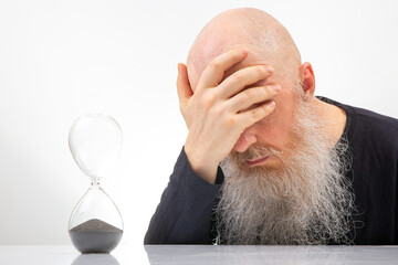 bald man with a large white beard covered his face with his hand in regret at the end of time in an hourglass. the concept of the transience of time. end of time measurement