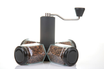 black manual coffee grinder for coffee beans and four glass transparent jars with black lids filled with roasted coffee beans and earthenware for brewing coffee on a white background. 