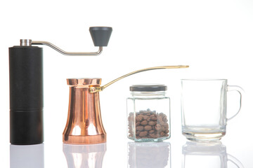 Close-up of a sleek black coffee grinder, a shiny copper cezve, a jar of coffee beans, and a transparent glass mug on a white surface, perfect for coffee preparation visuals.