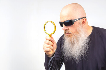 bearded man in sunglasses looks through a magnifying glass on a light background. search and research concept. detective agency