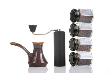 black manual coffee grinder for coffee beans and four glass transparent jars with black lids filled with roasted coffee beans and earthenware for brewing coffee on a white background. 
