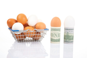 White and brown eggs in a metal basket next to rolled one- and two-dollar bills with an egg, set against a glossy white background for a financial food theme.