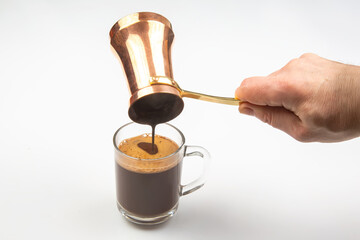 A hand pours Turkish coffee from a traditional copper cezve into a clear glass cup, showcasing the rich, frothy texture against a white background.