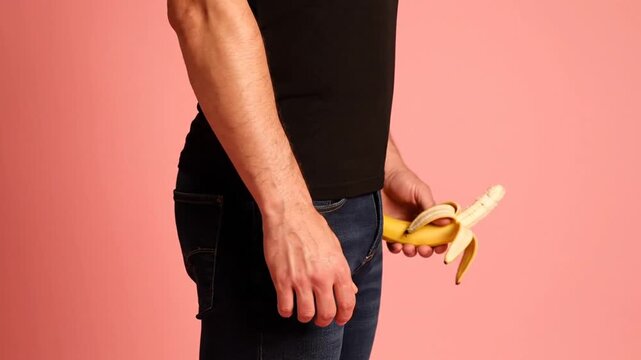 Man in Black T-Shirt and Blue Jeans Holding a Peeled Banana Against a Soft Pink Background