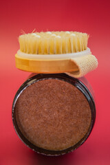 Natural body scrub jar with wooden lid and bristle brush on top, showcasing rich brown texture against a vibrant red background, emphasizing skincare and wellness