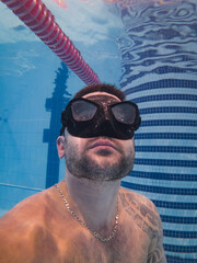 Close-up underwater portrait of a tattooed male freediver wearing a black mask training breath-hold in an indoor swimming pool.
