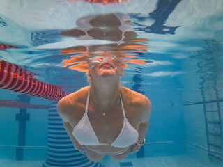 Underwater close-up view of a woman in a white bikini surfacing in a blue indoor swimming pool with water reflections.