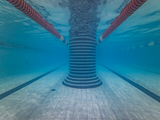 Symmetrical underwater view of an empty indoor swimming pool with a tiled central pillar and floating red lane dividers.