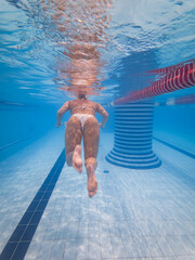 Rear view of a female swimmer in a white bikini swimming underwater in a blue tiled indoor pool.
