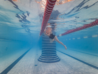 Underwater wide angle view of a female swimmer in a black swimsuit holding a lane rope in an indoor pool.