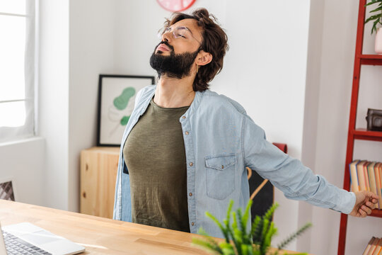 Young hipster freelancer man leaning back in chair doing stretching exercise resting at workplace with hands behind head after successful work. Business and home office concept.