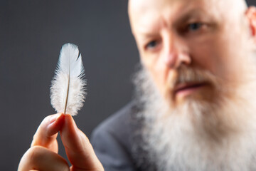an aged man with a full white beard focuses on a single delicate feather, held tenderly in his hand, representing themes of wisdom, gentleness, and the fragility of life in a contemplative setting