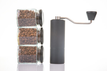 black manual coffee grinder for coffee beans and three glass transparent jars with black lids filled with roasted coffee beans and earthenware for brewing coffee on a white background. 
