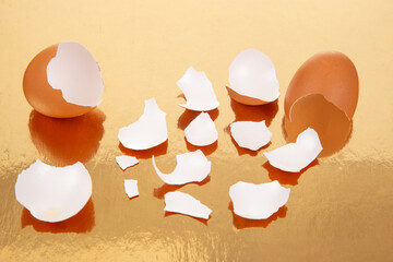 Two broken brown eggs with scattered eggshell fragments on a reflective golden surface, showcasing a dramatic and textured food photography scene with warm tones.