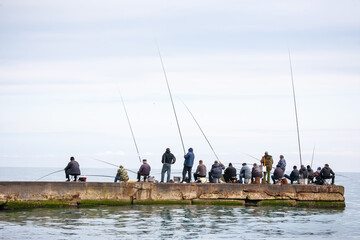 A group of people fishing with rods along a concrete pier by the sea under a cloudy sky. Some sit on chairs with gear, others stand, enjoying a peaceful day by the water.